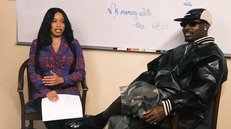 Ime Ekpo and the artist Asake are seated in chairs during an interview or conversation. Ime Ekpo, on the left, wears a blue and red patterned blazer and holds a stack of papers while looking toward Asake. Asake, on the right, wears sunglasses, a black leather jacket, a baseball cap, and camouflage shorts. Behind them is a whiteboard featuring the handwritten phrase, "Mr. money with the VIBE."