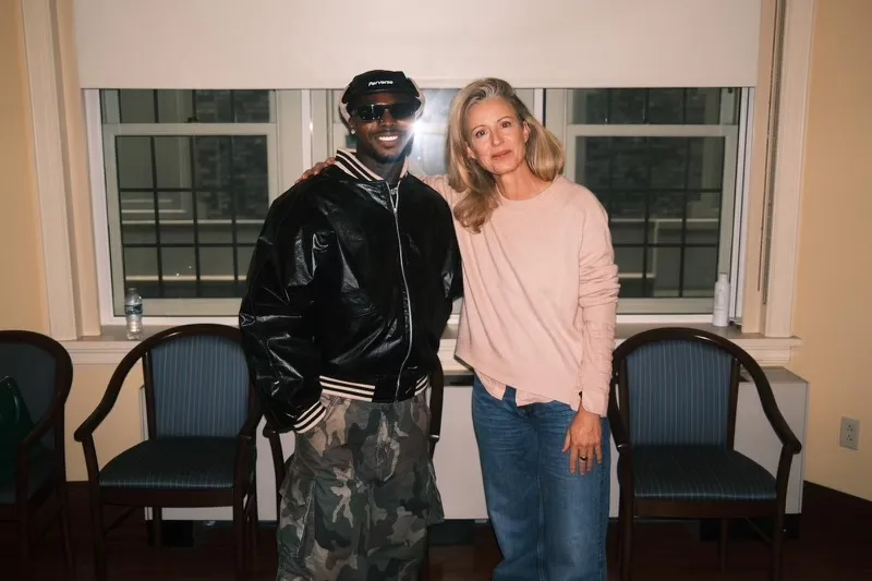 Associate Director of the Institute of African Studies (IAS) Jinny Prais stands with Afrobeats artist Asake in a room with large windows and several chairs. Jinny Prais, on the right, is wearing a light pink sweater and blue jeans. Asake, on the left, wears a black leather bomber jacket, camouflage cargo pants, a black baseball cap, and sunglasses.